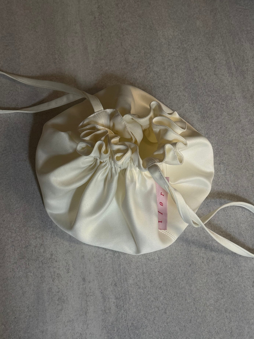 Bloom Pouch in ivory satin photographed from above as a flat lay, showing gathered ruffle detail and crossbody strap extended on both sides. 