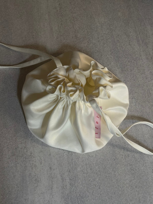 Bloom Pouch in ivory satin photographed from above as a flat lay, showing gathered ruffle detail and crossbody strap extended on both sides. 