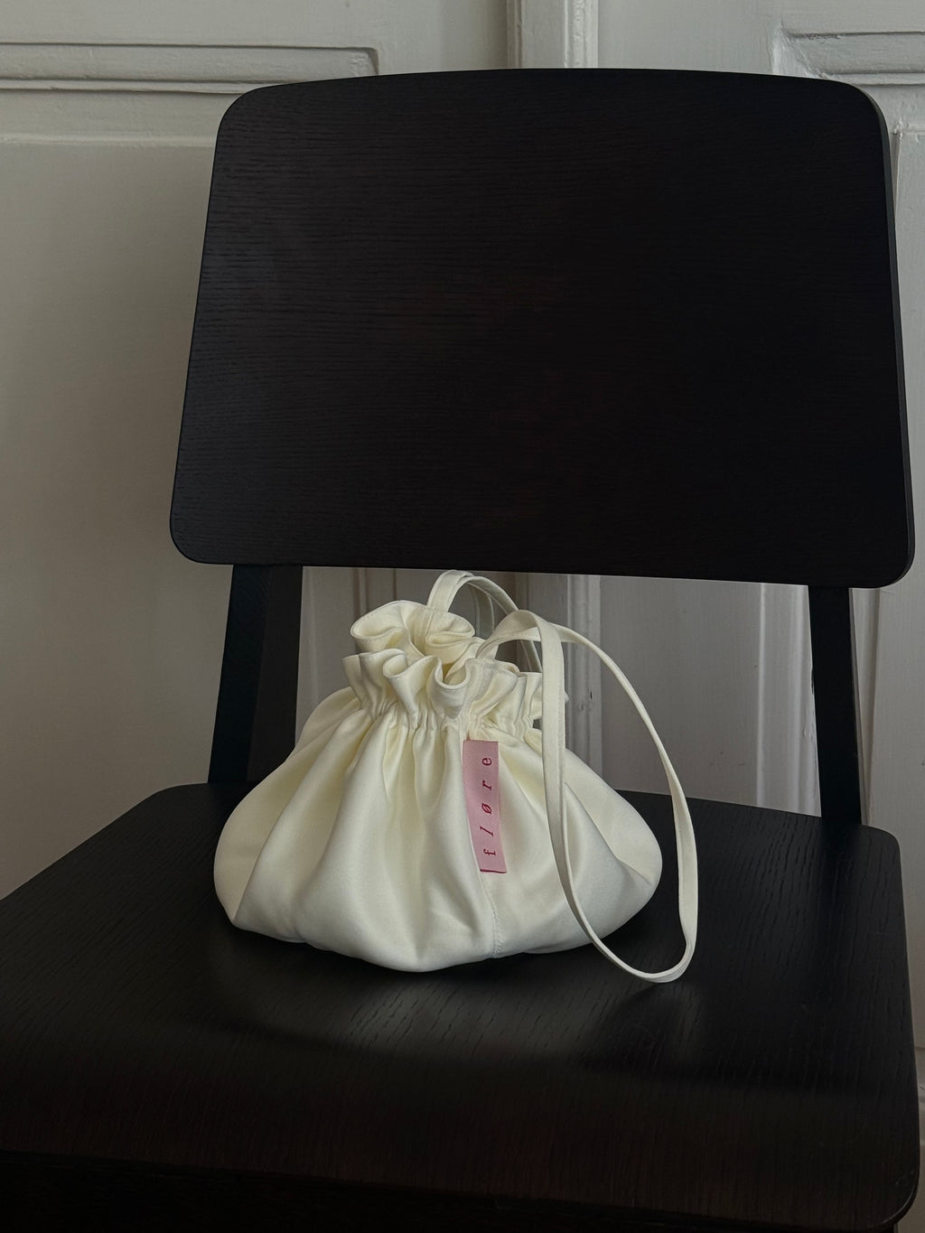 
Bloom Pouch in ivory satin sitting upright on a dark wooden chair, showing full rounded silhouette, ruffled drawstring opening, and Fløre label.