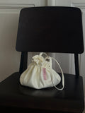 
Bloom Pouch in ivory satin sitting upright on a dark wooden chair, showing full rounded silhouette, ruffled drawstring opening, and Fløre label.