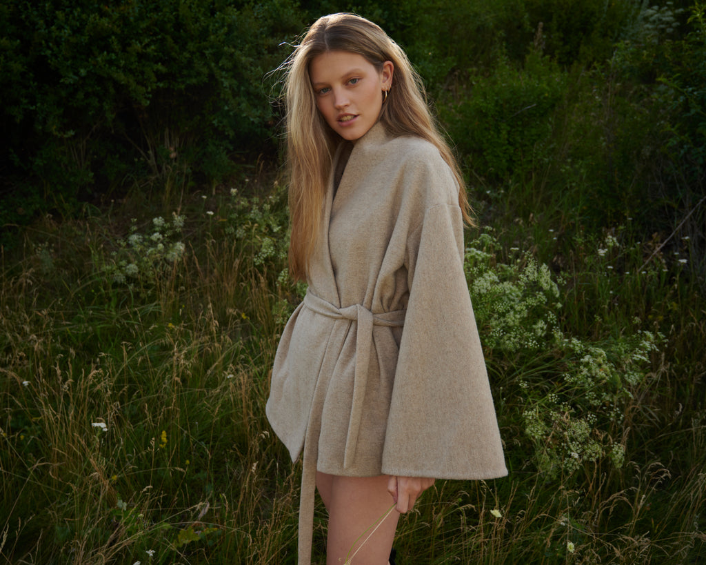 A woman wearing a light beige wool wrap coat with wide flared sleeves and a matching waist tie, standing in a grassy outdoor field.