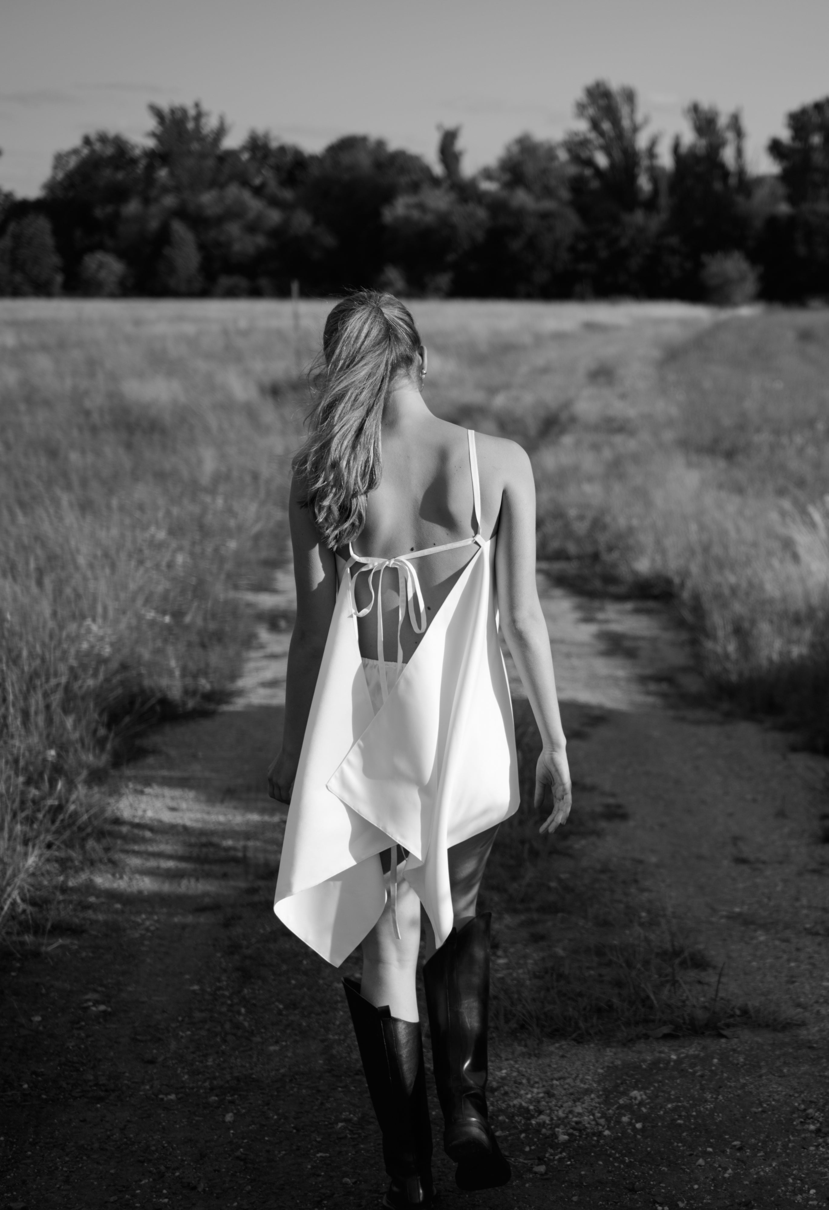 A woman viewed from behind walking on a dirt path through a grassy field, wearing a white backless wrap dress with tie straps and dark knee-high boots.