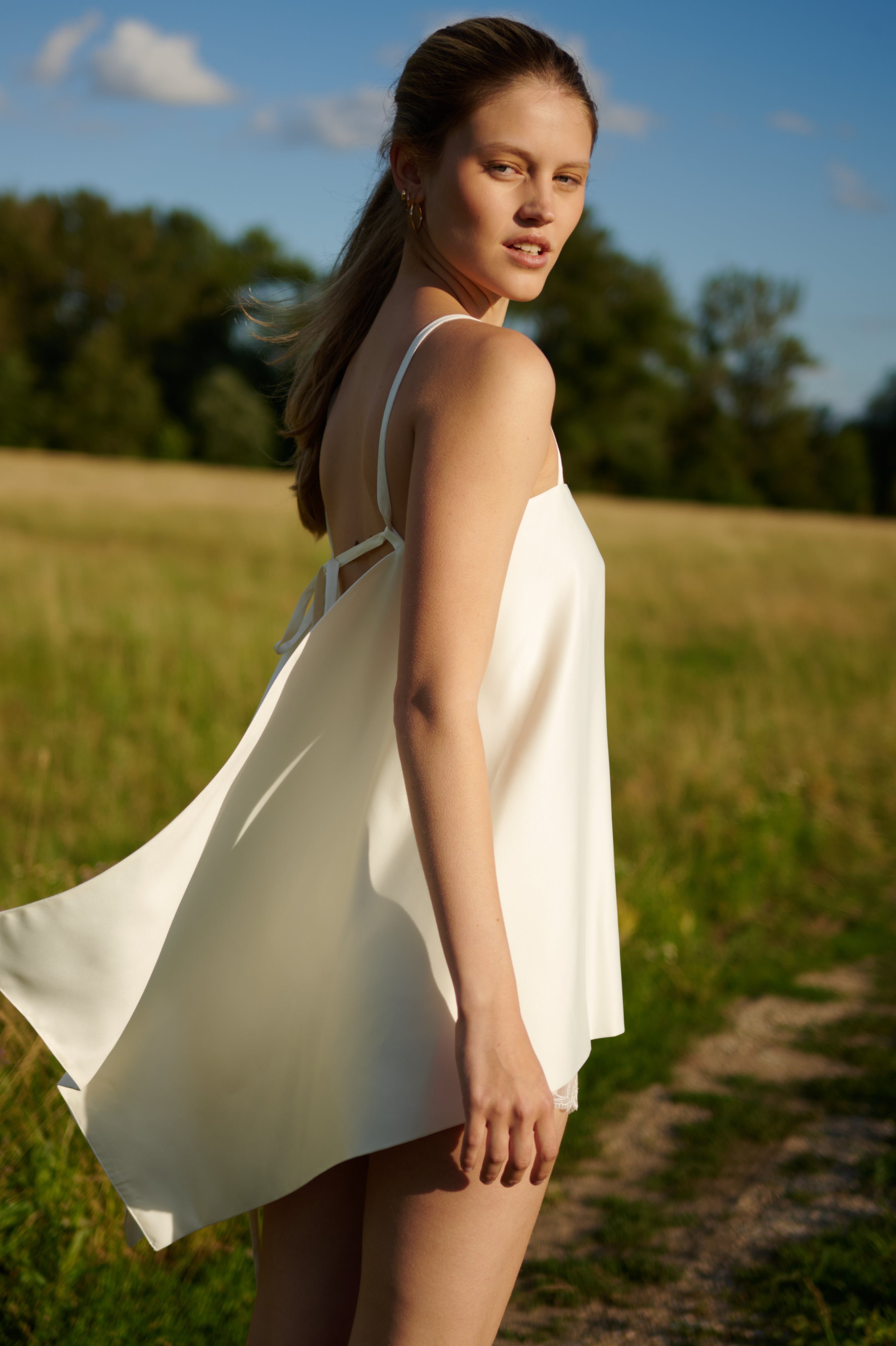A woman wearing a white sleeveless slip dress stands outdoors in a grassy field, looking back over her shoulder under a blue sky.