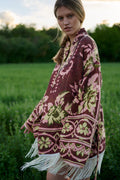 A woman wearing a bohemian-style patterned poncho with white fringe, standing in a grassy field at sunset.