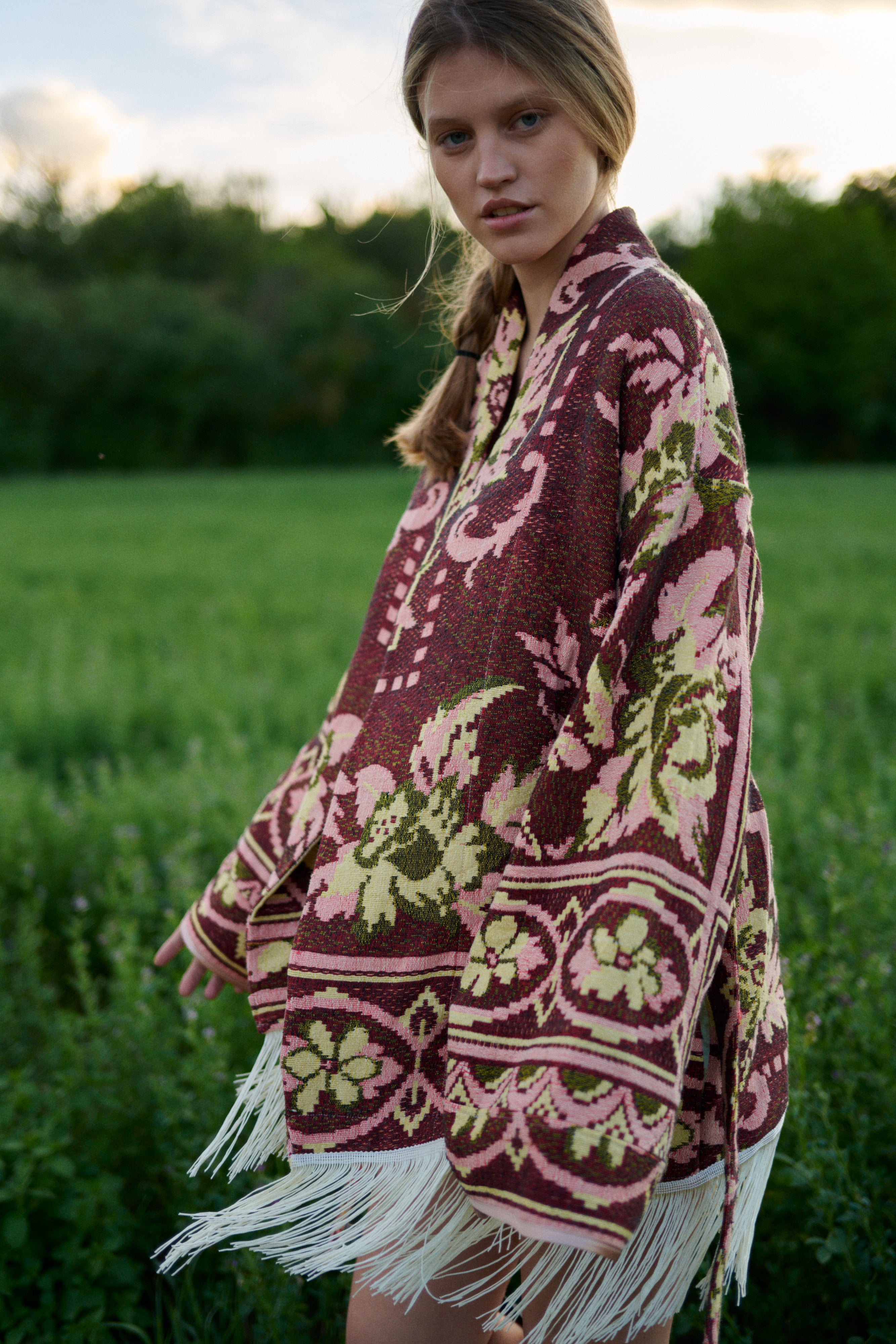 A woman wearing a bohemian-style patterned poncho with white fringe, standing in a grassy field at sunset.
